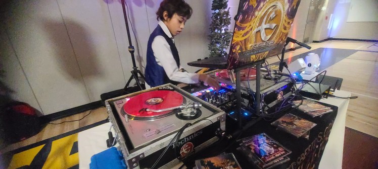 DJ Emerson of Denver's Best DJs in a vest and dress shirt Deejaying for The Coyote Hills Elementary School Winter Snowball Dance Party at Infinity Middle School. Technics Turntables with red serato records laptop with DJ Emir's DJEMIR.com Logo Christmas Trees in the background and DJ Emir Mixtapes on the table
